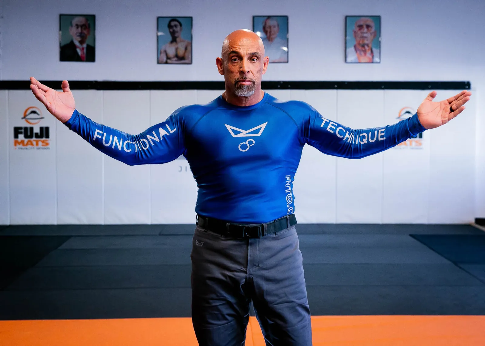 A man in the blue long sleeve FNTQ rashguard, arms out on the mats, FUNCTIONAL and TECHNIQUE down the sleeves